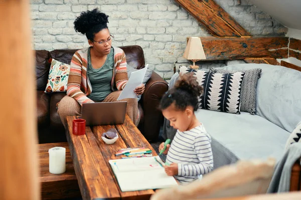African American stay at home mother working on laptop and analyzing documents while her daughter is drawing on the paper. 