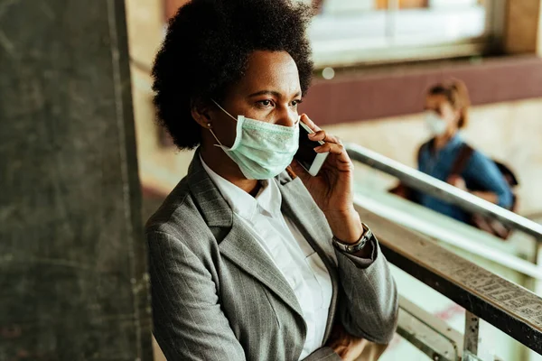 African American businesswoman with protective face mask communicating over smart phone while being at public hallway at railroad station. 