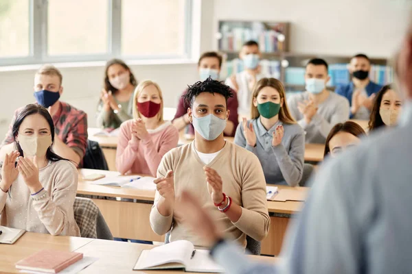 Happy university students applauding after successful lecture while wearing protective face masks due to COVID-19 pandemic. Focus is on black male student.