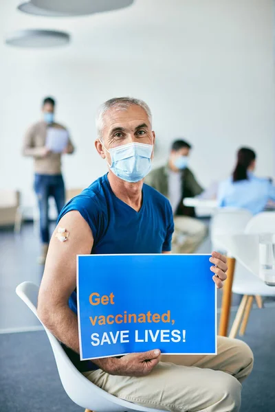 Mature man with face mask holding placard after receiving COVID-19 vaccine and inviting people to get vaccinated and save lives.