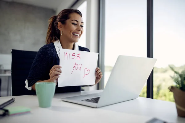 Happy businesswoman making video call over laptop and holding a placard with \'Miss you\' inscription.