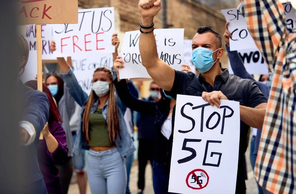 Caucasian man wearing protective face mask and holding a banner with Stop 5G inscription while protesting with crowd of people on city streets.