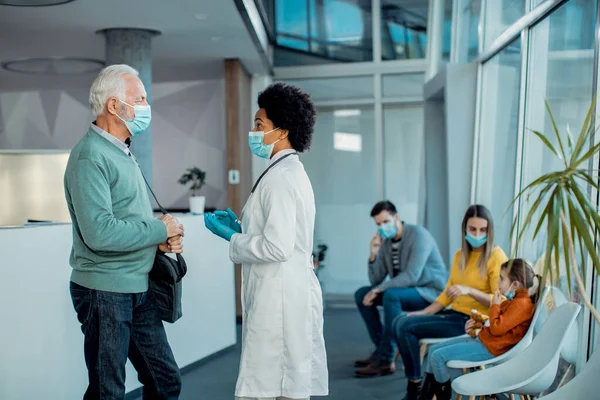 Black female doctor communicating with senior patient while standing in a lobby at the hospital. They are wearing face masks due to coronavirus pandemic.