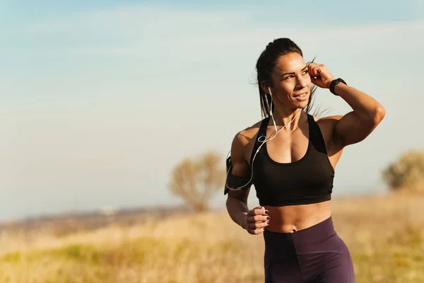 Young muscular build woman adjusting earphones while running in nature. Copy space.