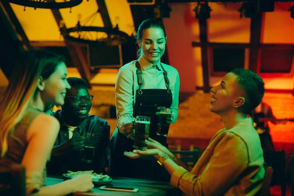 Happy waitress serving drinking to group of young people while working at night in a bar.