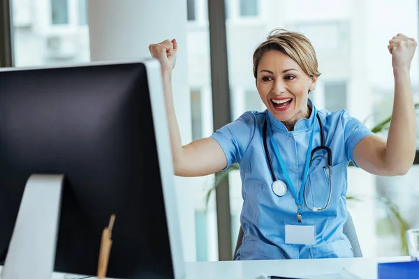 Happy female healthcare worker reading an e-mail on desktop PC and celebrating good news at medical clinic.