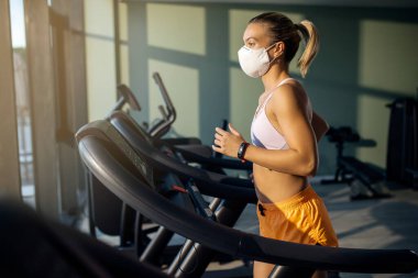 Dedicated female athlete jogging on running track while wearing protective face mask in a gym during coronavirus epidemic. 
