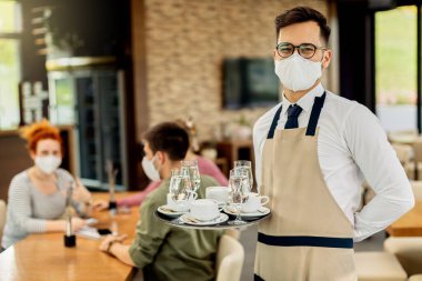 Happy waiter wearing protective face mask while serving coffee to guests during coronavirus epidemic. 