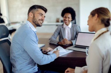 Happy man talking to his wife while signing contract with insurance agent during the meeting in the office. 