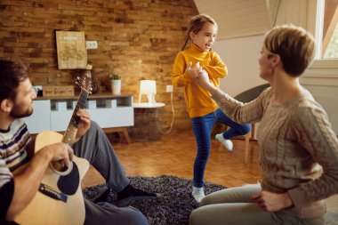 Happy little girl having fun and dancing while her father is playing acoustic guitar at home.