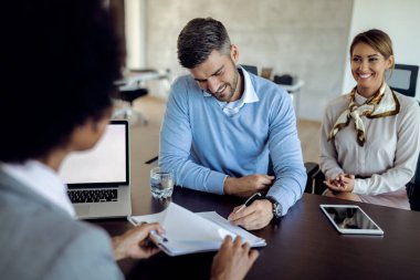 Young happy man signing a contract while being with his wife on a meeting with their bank manager. 