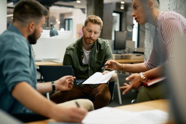Freelance worker using laptop while going through paperwork with his colleagues at corporate office.