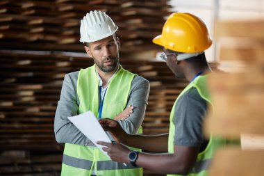 Warehouse manager talking while analyzing paperwork with male worker at lumber department. 
