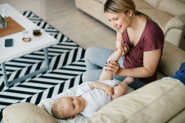 Cute baby boy lying down on sofa while mother is massaging his leg at home.