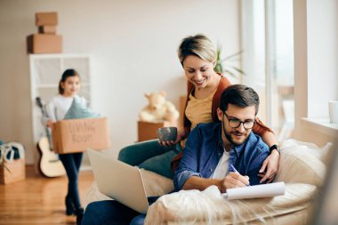 Happy man using laptop and going through paperwork while moving into a new home with his wife and daughter. 