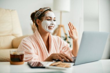 Young happy woman using laptop and waving to someone during video call while wearing white cosmetic face mask.