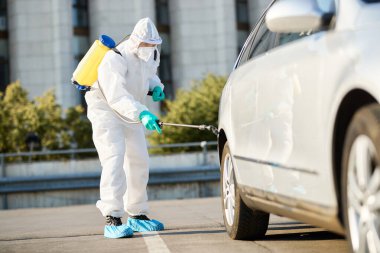 Man in protective hazmat suit disinfecting car tire during coronavirus pandemic. 