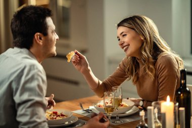 Happy woman having fun while feeding her boyfriend during dinner at dining table. 
