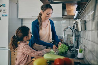 Happy mother and her small daughter washing vegetables under kitchen sink while preparing food together at home. 
