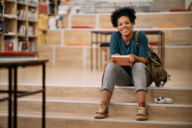 Happy African American female student relaxing on staircase in library and looking at camera.