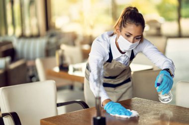 Young waitress with face mask disinfecting restaurant tables due to coronavirus pandemic.