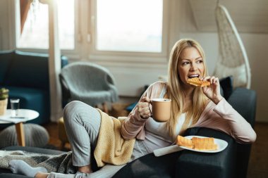 Young woman drinking coffee while siting on the sofa and eating waffles in the living room. 