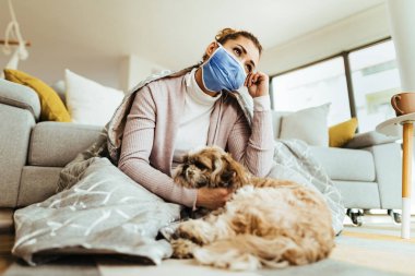 Low angle view of pensive woman thinking while wearing protective face mask and sitting on the floor with her dog at home. 