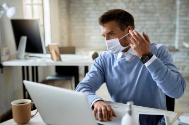 Businessman putting on protective face mask while working on laptop in the office. 