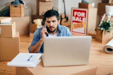 Displeased man talking on the phone and using laptop while relocating in a new apartment. 