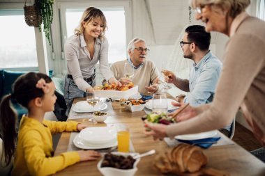 Happy multi-generation family having lunch together at home. Men are talking among themselves while women are serving food at dining table. 