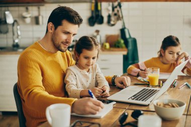 Little girl sitting on father's lap while he is working on laptop and writing notes at home. 