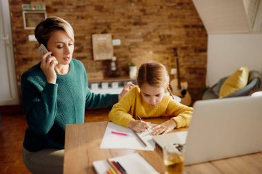 Single mother talking on mobile phone while her daughter is drawing on the paper at home. 