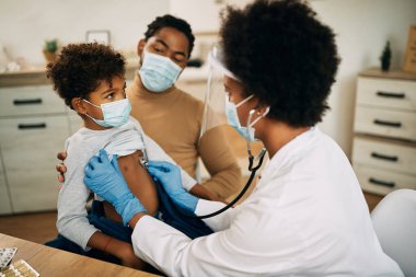 African American boy with face mask being examined by female pediatrician during medical apportionment at doctor's office. 