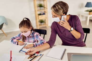 Young mother working at home and talking on the phone while homeschooling her daughter due to coronavirus pandemic.