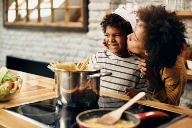 Loving African American mother kissing her small son while cooking together in the kitchen. Focus is on boy.