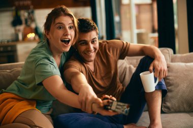 Young woman watching TV in disbelief while boyfriend is trying to take remote control from her at home.