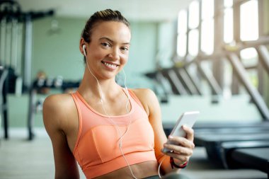 Happy athletic woman listening music on earphones while using smart phone in a gym.