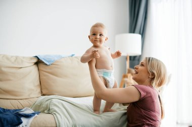 Cute boy standing on the sofa with help of his happy mother at home.