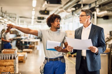 Mid adult manager and black worker analyzing project plans while walking through industrial facility. 