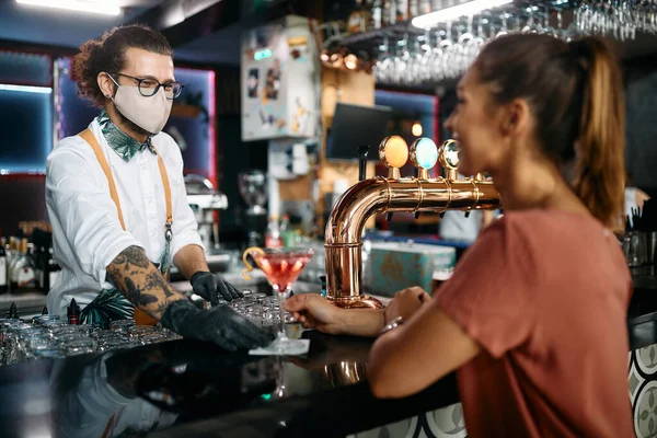 Happy bartender with face mask serving cocktail to a woman at bar counter.