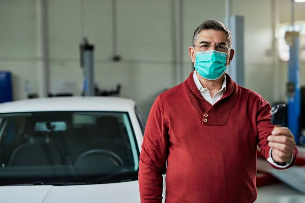 Male customer with protective face mask holding a key of his repaired car in a workshop and looking at camera.