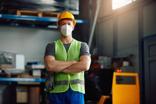 Portrait of warehouse worker with arms crossed wearing face mask at work due to coronavirus pandemic.