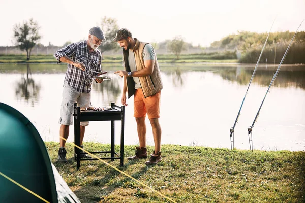 Happy man and his senior father preparing meat on barbecue grill while camping by the river in nature. Copy space.