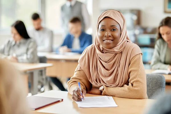 Happy African American female student writing an exam in the classroom and looking at camera.