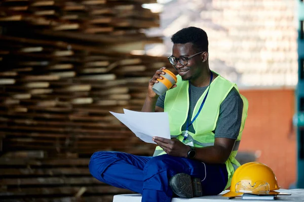 Young African American worker going through reports during his coffee break at wood warehouse.