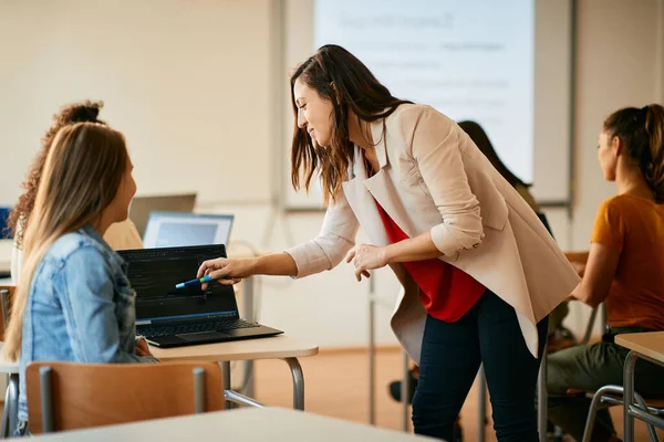 Smiling IT teacher helping her student in learning computer language on laptop during high school class.