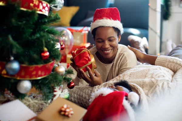 Happy black woman holding a gift while talking to her husband by Christmas tree at home.