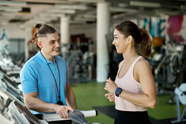 Happy female athlete talking with personal trainer while running on treadmill and working out in a gym.