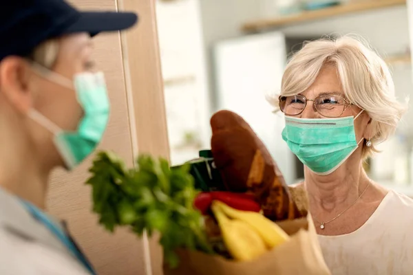 Happy mature woman wearing protective face mask while talking to female courier who is delivering hr groceries during COVID-19 quarantine.