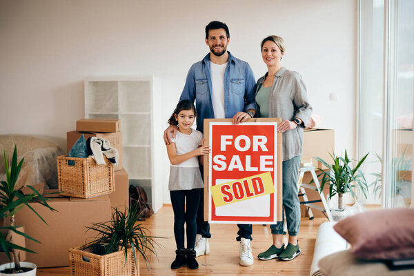 Happy family standing in their new home while holding sold real estate sing and looking at camera. 